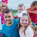 Um grupo multiétnico de crianças vestindo roupas coloridas brinca ao ar livre. No primeiro plano, um menino de pele clara abraça uma menina com síndrome de Down que usa uma laço rosa no cabelo. Ambos sorriem