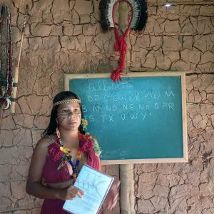 Na foto, uma mulher indígena leciona uma aula. Ela está na frente de uma lousa, que contém letras do alfabeto tupi-guarani.