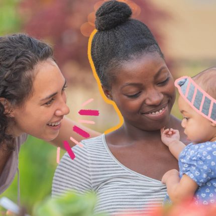Duas mulheres interagem com um bebê. A mulher que segura a criança no colo é negra e usa camiseta branca. A outra mulher tem pele clara e cabelos crespos. O bebê usa uma roupinha azul e tiara.