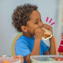 Na imagem, um menino negro come um sanduíche. A imagem possui intervenções de rabiscos coloridos.