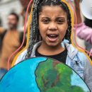 Na imagem, uma menina negra está em um protesto. Na frente dela, há o desenho de uma Terra. A imagem possui intervenções de rabiscos coloridos.