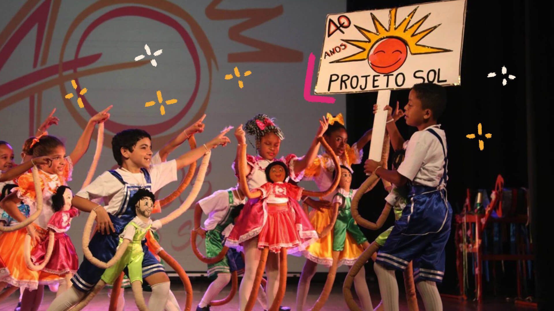 Na foto, um grupo multiétnico de crianças dança em um palco. Na frente, um menino negro segura uma placa escrito 40 anos - Projeto Sol, com o desenho de um sol sorridente. A imagem possui intervenções de rabiscos coloridos.