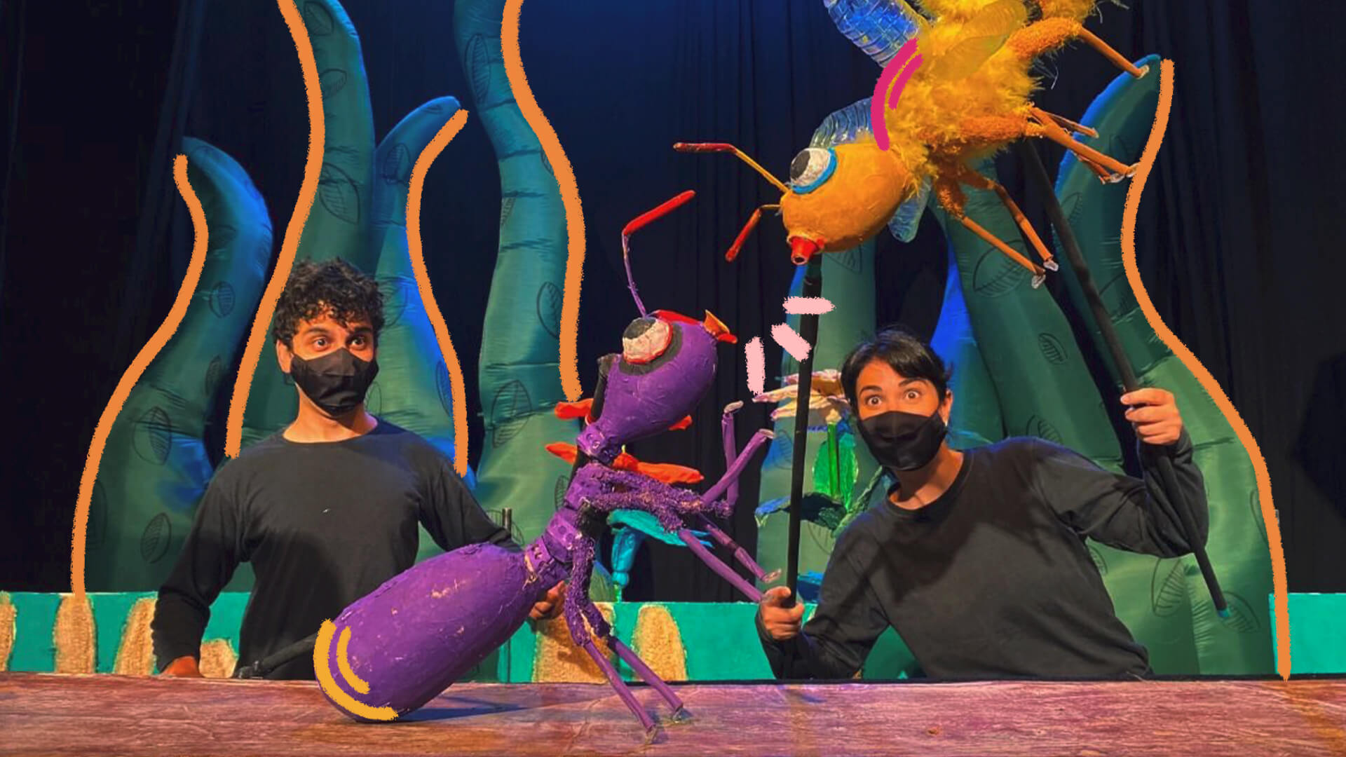 Na foto, um ator e uma atriz manipulam um boneco de uma formiga na cor roxa e de uma abelha amarela. Os atores tem tonalidade de pele alaranjada devido à iluminação do cenário.