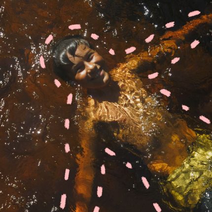 Na foto, uma criança de pele escura aparece sorrindo enquanto brinca mergulhada em um rio. a imagem possui intervenções de rabiscos coloridos na cor rosa.