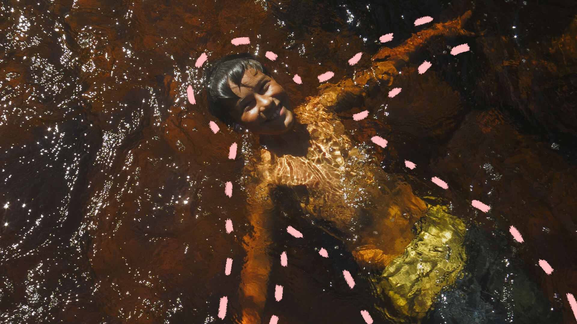 Na foto, uma criança de pele escura aparece sorrindo enquanto brinca mergulhada em um rio. a imagem possui intervenções de rabiscos coloridos na cor rosa.