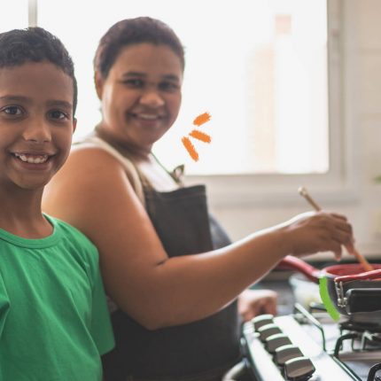 Na imagem, mãe e filho estão cozinhando em uma cozinha. Ambos sorriem e são negros. A imagem possui intervenções de rabiscos coloridos.