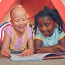 Duas meninas leem um livro embaixo de uma cabaninha. Uma delas é uma criança branca sem cabelo que tem alopecia universal e a outra é uma menina negra que usa tranças no cabelo
