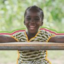 Na foto, uma menina negra sorri enquanto se apoia em uma mesa de madeira. A imagem possui intervenções de rabiscos coloridos.