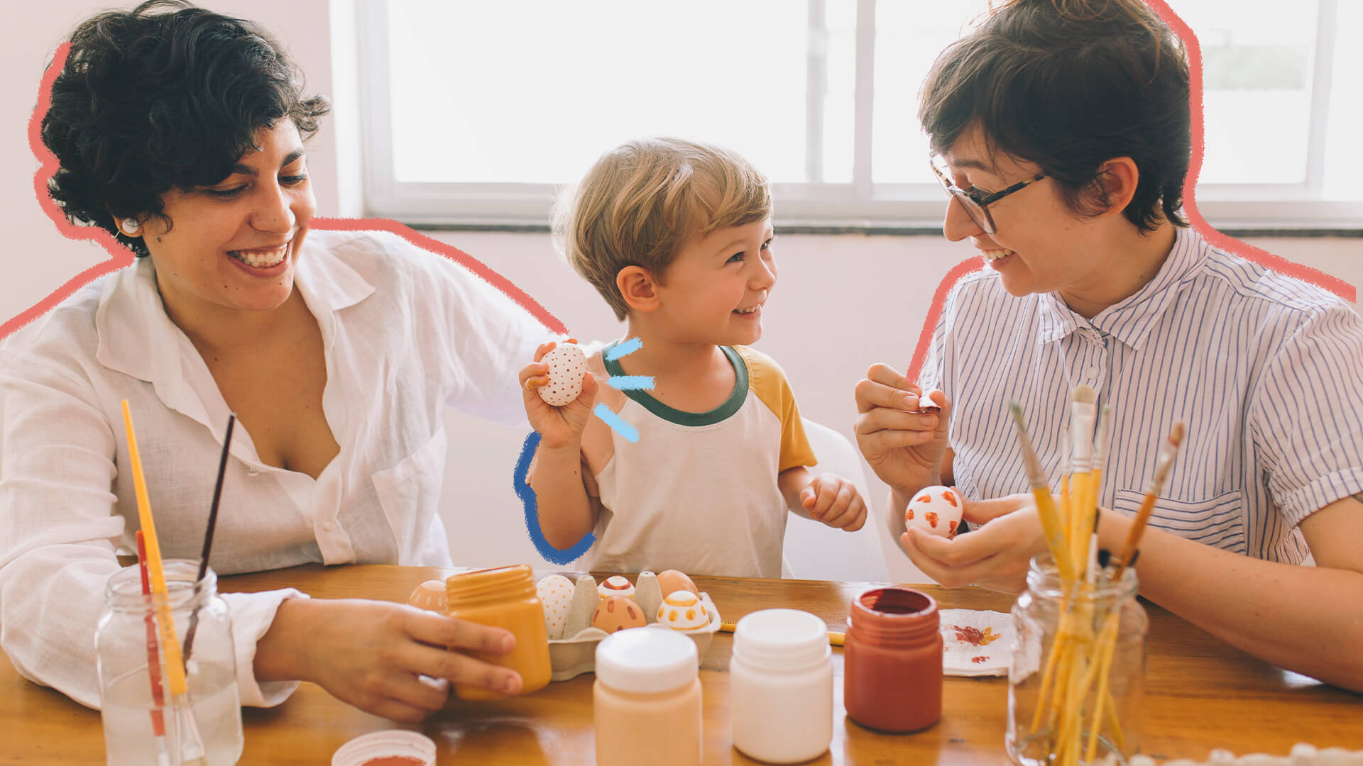 Feriado de páscoa: na foto, uma família de duas mamães e um menino pintam ovos para a páscoa. Elas têm pele branca e cabelos escuros, enquanto o menino é loiro. Todos sorriem. A imagem possui intervenções de rabiscos coloridos.