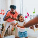 Uma família de pessoas negras (dois adultos e duas meninas) dançam na sala de casa