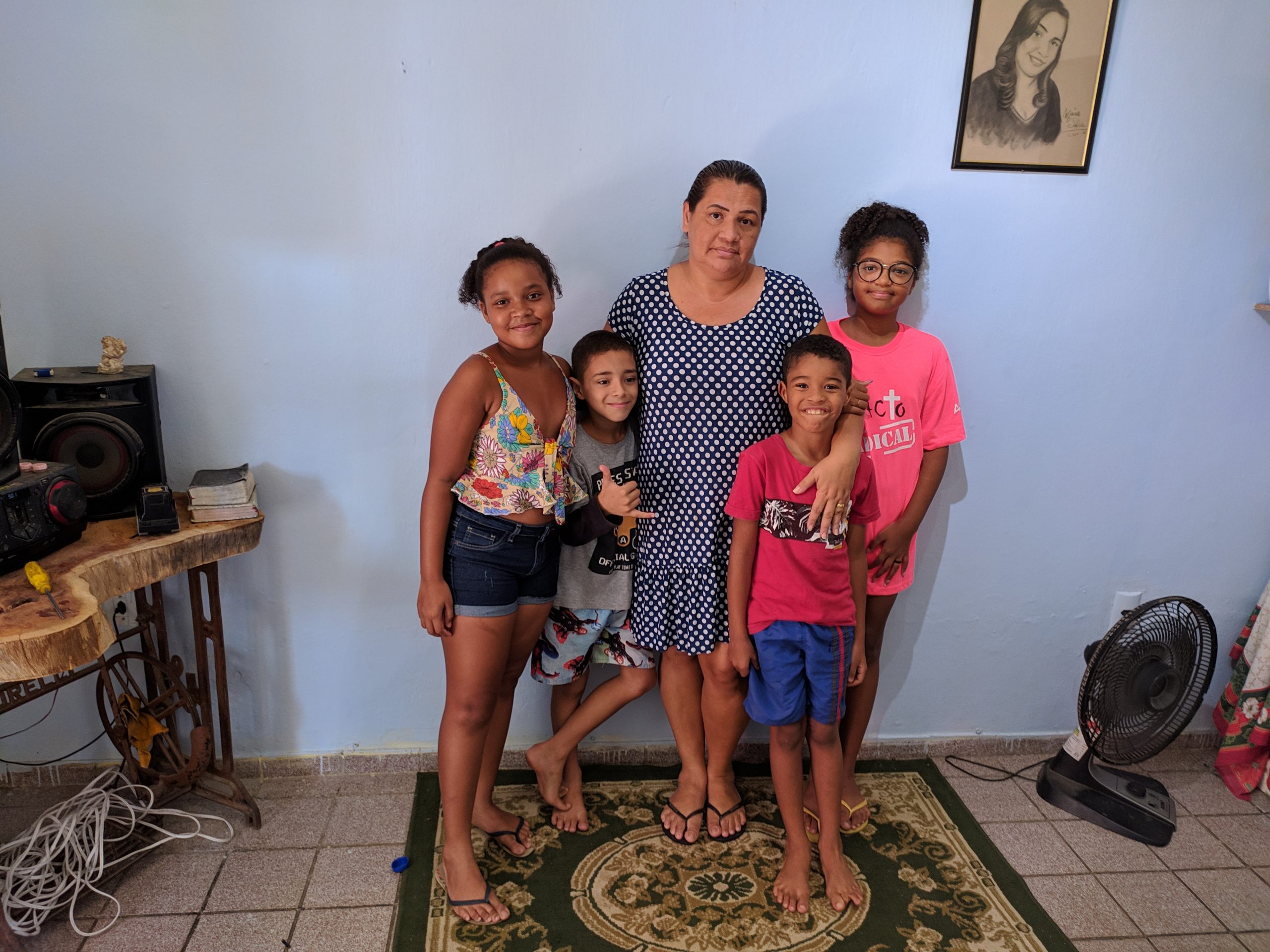 Uma mulher adulta e quatro crianças posam para a foto na sala de casa. Elas estão posicionadas em cima de um tapete; atrás, há uma parede branca e um quadro