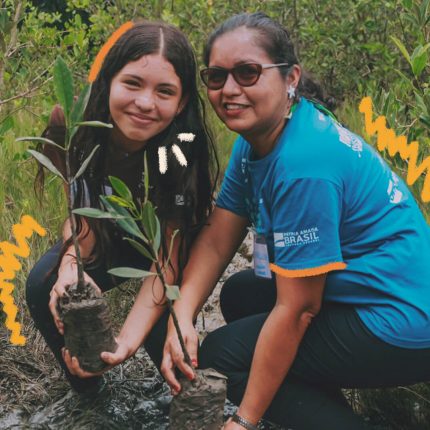 Uma menina e uma mulher adulta olham para a foto enquanto seguram mudas de árvores. As duas estão agachadas numa área de mangue.