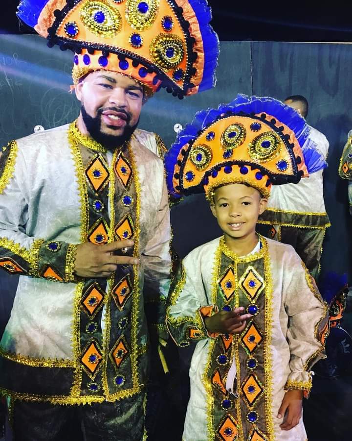 Um homem e um menino, negros, estão fantasiados e prontos para desfilar na escola de samba.