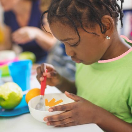 Uma menina negra está sentada à mesa olhando para a comida em seu potinho entre as mãos.