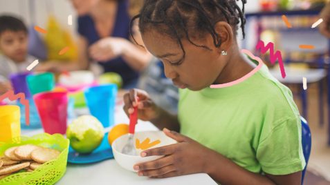 Uma menina negra está sentada à mesa olhando para a comida em seu potinho entre as mãos.