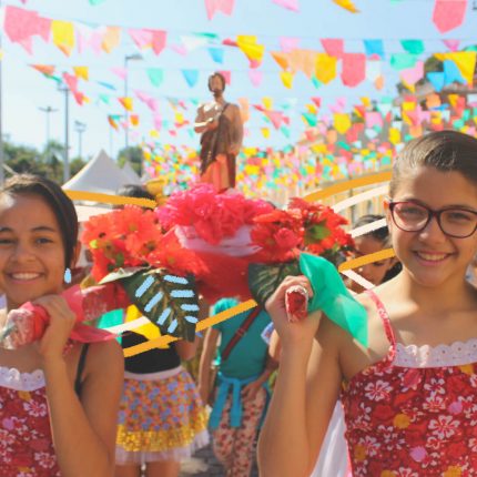 Duas meninas de vestidos floridos em vermelho iguais seguram o andor de São João ornamentado com flores vermelhas