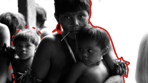 A foto mostra uma mulher indígena de cabelos curtos e brincos segurando um menino indígena contra o peito.