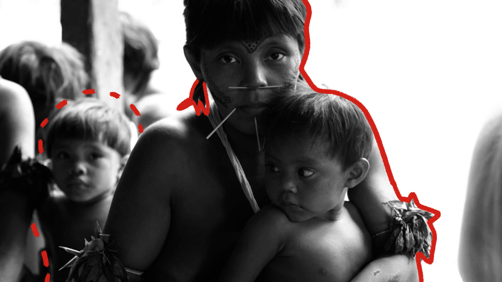 A foto mostra uma mulher indígena de cabelos curtos e brincos segurando um menino indígena contra o peito.