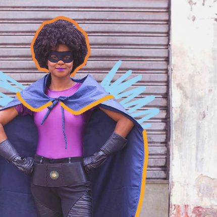 Na foto, uma mulher negra veste uma roupa de super herói na cor roxa. Ela está na frente de uma porta de metal. A imagem possui intervenções de rabiscos coloridos.