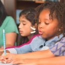 Uma turma de alunas participa de uma aula. Em destaque, uma menina negra escreve com um lápis em uma folha em branco.