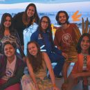 Jovens que participaram do documentário "Educação, presente para o futuro" posam ao lado da diretora Patricia Travassos Todos usam roupas coloridas e o fundo é azul