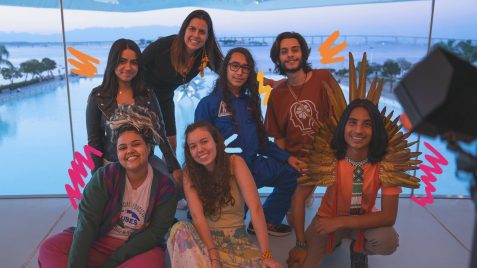 Jovens que participaram do documentário "Educação, presente para o futuro" posam ao lado da diretora Patricia Travassos Todos usam roupas coloridas e o fundo é azul