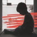 Imagem em preto e branco de um menino sentado no chão, olhando para baixo. A foto mostra sua silhueta contra a luz. A imagem possui intervenções de rabiscos vermelhos.