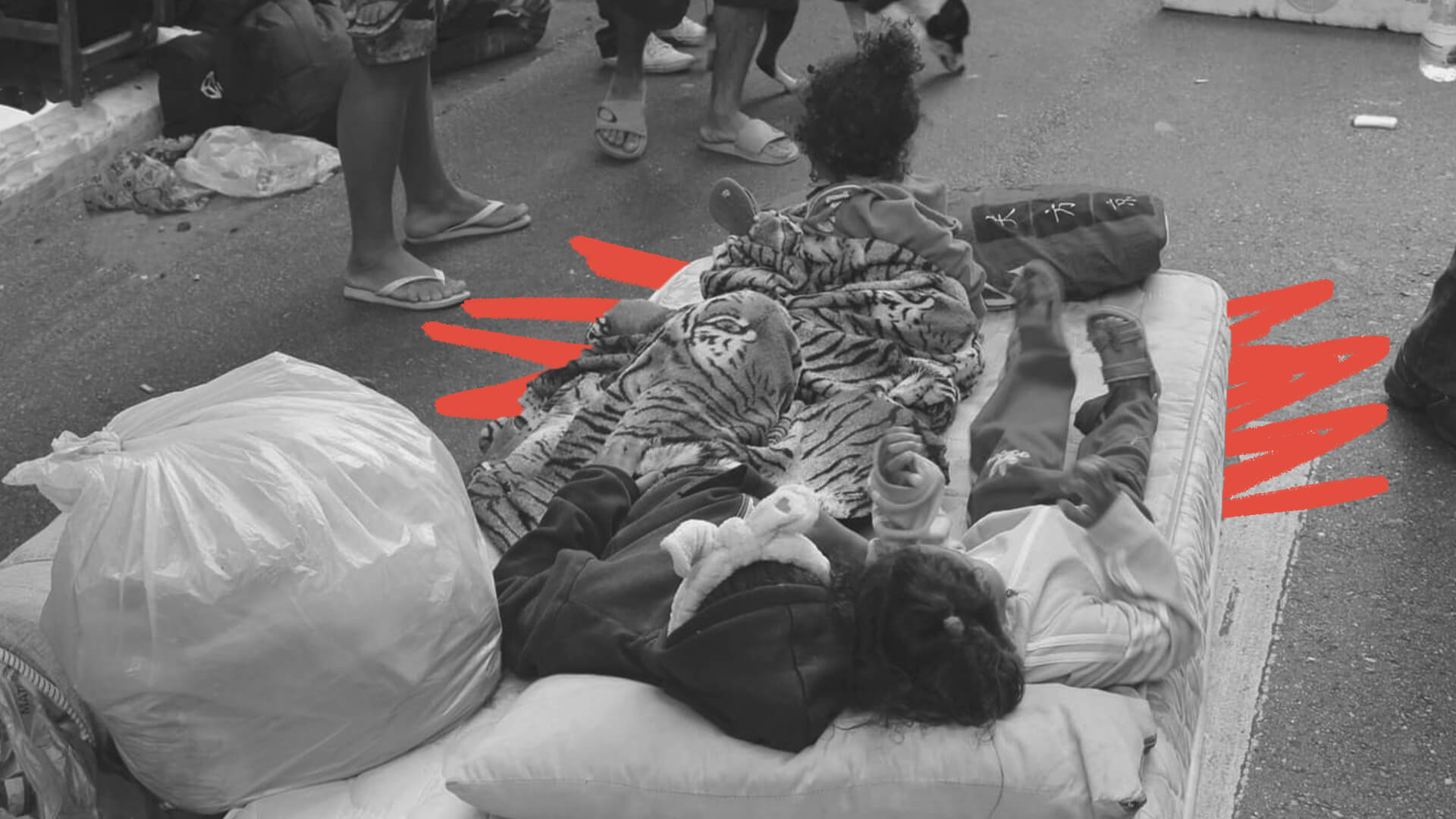 Foto em preto e branco com duas crianças deitadas num colchão, na rua, sem mostrar seus rostos. Ao redor delas, há um saco plástico e pés de outras pessoas
