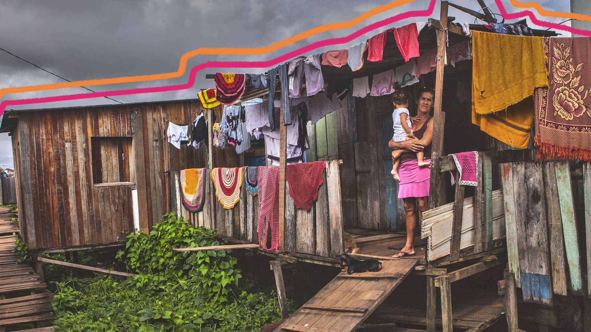 Na imagem, uma mulher segura uma criança no colo. Elas estão em frente à porta de uma casa de madeira precária, com roupas coloridas estendidas no varal.