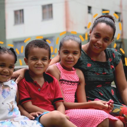 Uma família negra está sentada em um banco, na parte externa de um condomínio. A família é composta por uma mulher e três crianças, sendo elas um menino e duas meninas.