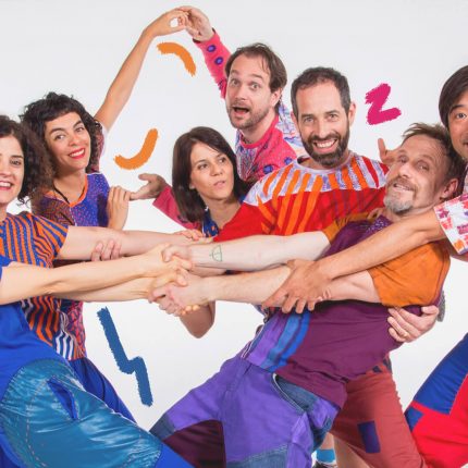 Foto do grupo Barbatuques: homens e mulheres vestem roupas coloridas, alguns se dão as mãos e posam com movimentos de corpo diferentes
