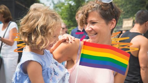 Coletivos LGBTQIA+: Uma mãe segura a filha no colo junto de uma bandeira de arco-íris.