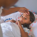 Uma menina de pele bronzeada está deitada em uma cama. Uma mulher afere sua temperatura com a mão na testa. A imagem possui intervenções de rabiscos coloridos.