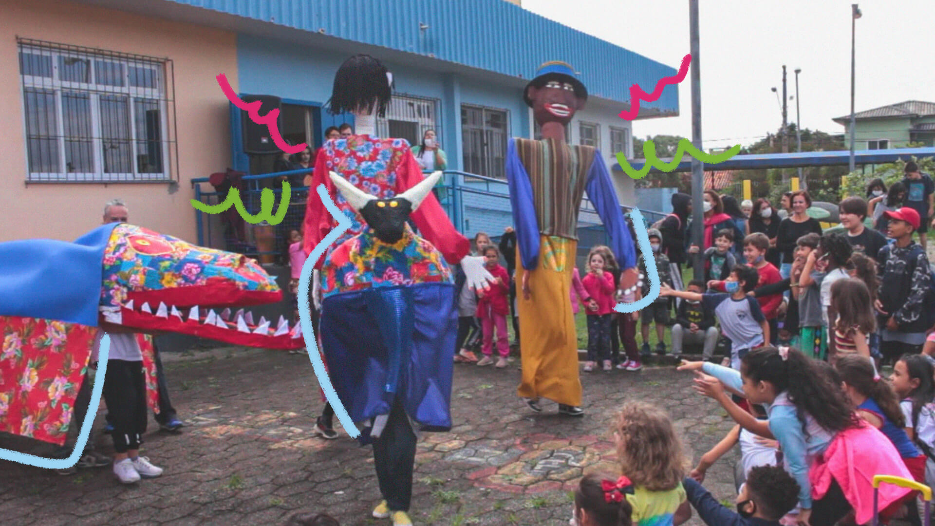 Dois personagens do folguedo Boi de mamão com roupas coloridas se apresentam no pátio de uma escola e as crianças assistem à volta