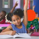 Na imagem, uma menina negra levanta o braço, se preparando para fazer uma pergunta na sala de aula. A imagem possui intervenções de rabiscos coloridos.