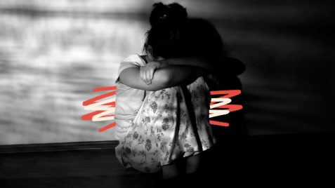 Foto em preto e branco de uma menina sentada no chão, que esconde o rosto o nos braços, apoiados no joelho. A imagem possui intervenções de rabiscos coloridos e ilustra uma matéria sobre revitimização.