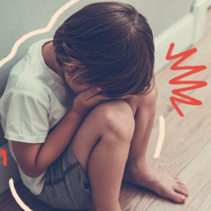 Um menino de pele clara está sentado no chão, escondendo a cabeça nas mãos e joelhos.