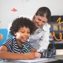 Na imagem, um menino negro sorridente escreve em um caderno. Atrás dele, uma professora branca também sorri enquanto olha a criança.