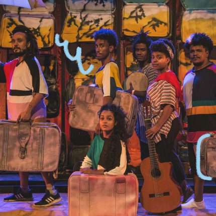 Um grupo de atores em um palco, vestindo roupas coloridas e com malas grandes nas mãos