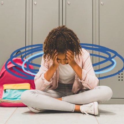 Uma menina negra está sentada no chão, com as pernas cruzadas e segurando a cabeça com as mãos. Atrás dela há armários na cor cinza.