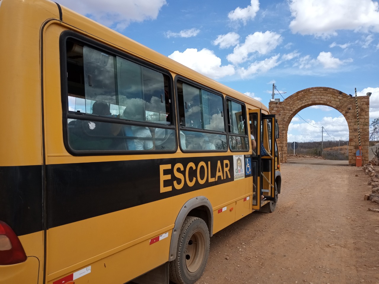 Um ônibus amarelo com uma faixa preta onde está escrito "Escolar" segue em direção ao portal de um parque em Canudos