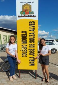 Duas meninas de camiseta branca do uniforme escolar posam junto de uma placa amarela onde se lê "Colégio modelo Maria José de Souza Alves"