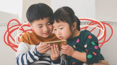 Duas crianças de etnia amarela olham para uma tela de celular.