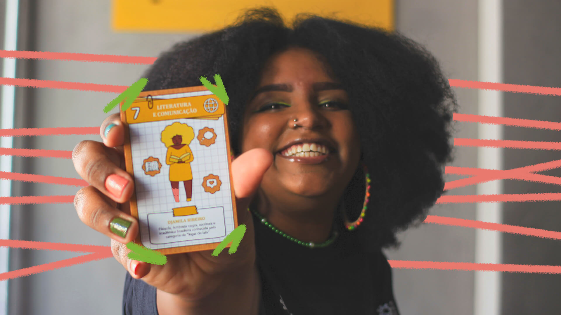 A foto mostra uma menina negra de cabelos crespos sorrindo e apresentando, em primeiro plano, uma carta do jogo Canindé, com um desenho de Djamila Ribeiro.