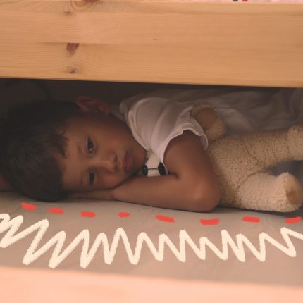 Uma criança está deitada no chão, escondida embaixo de uma cama.