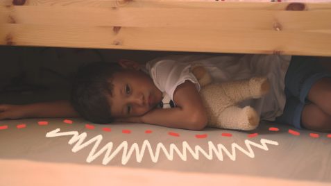 Uma criança está deitada no chão, escondida embaixo de uma cama.