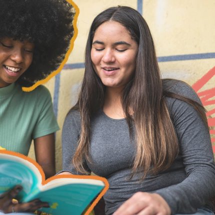 Na imagem, duas estudantes leem o mesmo livro. Uma é negra e a outra é branca.