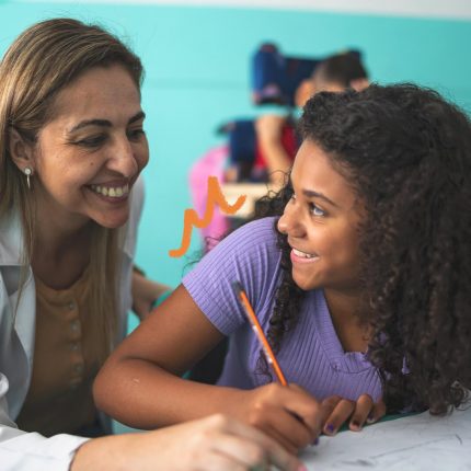 Uma menina negra sorri enquanto escreve em um caderno, acompanhada de uma professora, de pele clara, que também sorri.