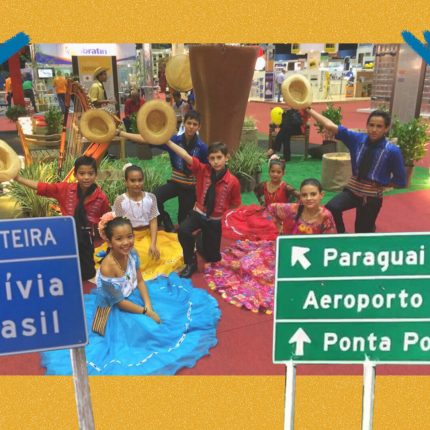 Foto de um grupo de crianças estão sentadas posando para uma foto no chão de um pavilhão com trajes coloridos de dança. Há duas placas; na azul, se lê Fronteira Bolívia Brasil; na verde, Paraguai Aeroporto Ponta Porã