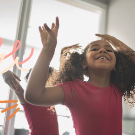 Duas crianças negras dançam em um ambiente interno. Na frente, uma menina negra de cabelos cacheados sorri.